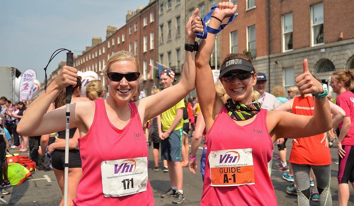Two smiling runners in pink race shirts give a thumbs up during a city race; one wears a Guide bib