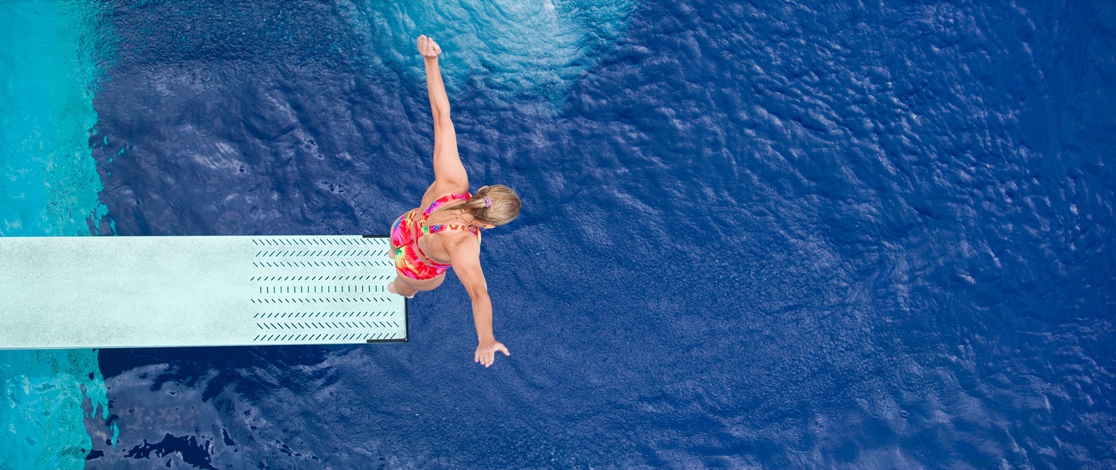 Seen from above, a diver leaps off a diving board toward deep blue water