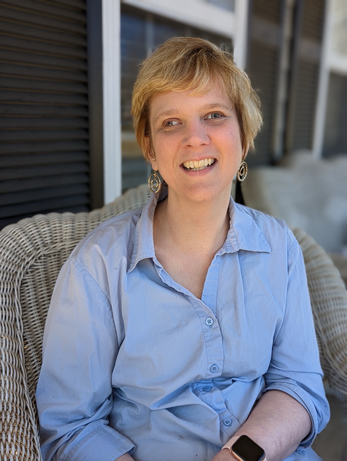 Smiling woman with short blonde hair in a light blue shirt, seated in a wicker chair on a porch