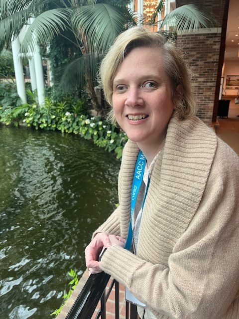 Smiling woman with short blonde hair in a cream cardigan, standing beside an indoor garden pond